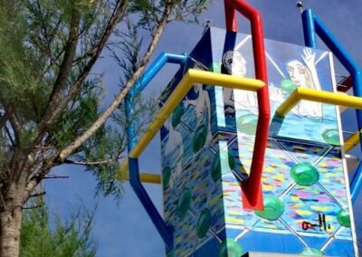 Intervención artística en la Torre Rhin-Maremondo Roberto Orallo | Santander, Cantabria La Torre Rhin-Maremondo, en Santander, es uno de los pocos ejemplos en la ciudad donde la arquitectura se fusiona con el arte público de la mano de un mismo autor: Roberto Orallo. Tanto el mural que viste el edificio como la escultura que lo acompaña en su base llevan la firma del artista, creando un diálogo permanente entre forma, color y memoria colectiva. El mural, con su lenguaje geométrico y sus colores vibrantes, transforma la fachada en una superficie dinámica, que responde al ritmo del entorno urbano y la luz cambiante del Cantábrico. Es una obra que invita a mirar hacia arriba, a descubrir el arte integrado en la ciudad. A sus pies, la escultura homenaje a los bañistas de la Primera Playa del Sardinero evoca la tradición veraniega de Santander. La pieza captura la esencia lúdica y popular de quienes han disfrutado del mar, el sol y la arena a lo largo de generaciones. Más que una figura literal, la obra es una reinterpretación poética de la experiencia del baño y el ocio junto al mar, traducida en formas estilizadas y evocadoras. Con esta doble intervención, Orallo celebra la identidad de Santander: su mar, su luz y su gente, en una conjunción de arte, paisaje y memoria.